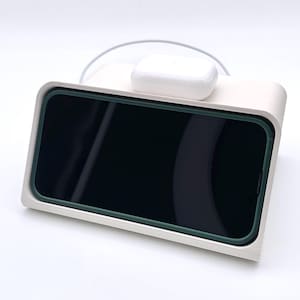 Può includere: Un supporto di ricarica bianco con un telefono nero e auricolari wireless bianchi in cima. Il supporto ha un bordo rialzato attorno al telefono e un piccolo labbro nella parte anteriore.