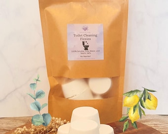 May include: A brown paper bag filled with white toilet cleaning fizzies. The bag has a label that reads "Toilet Cleaning Fizzies" and details about the product. Several fizzies are stacked in front of the bag, with a lemon and eucalyptus sprig in the background.