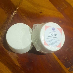 May include: Two white shower steamers, one unwrapped and one wrapped in clear plastic. The wrapped steamer has a pink label with the word "Love" and ingredient information. The background is a dark wooden surface.