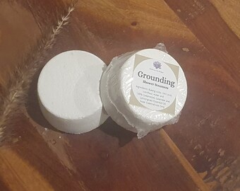 May include: Two white, round shower steamers labeled "Grounding" on a wooden surface. One steamer is unwrapped, and the other is wrapped in clear plastic. The label lists ingredients including baking soda, cornflour, and essential oils.