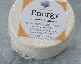 May include: A white, round shower steamer wrapped in clear plastic. The label reads "Energy" with ingredients listed, including essential oils of lemon, grapefruit, and orange. The product is designed for external use.