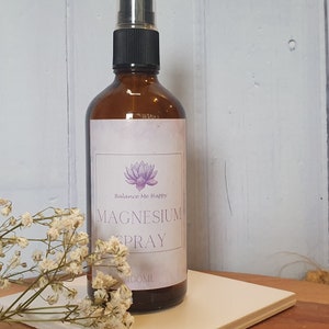 May include: A brown glass bottle with a black spray nozzle, labeled "Magnesium Spray" with a lotus flower illustration. The bottle contains 100ml of liquid. White flowers are in the foreground.