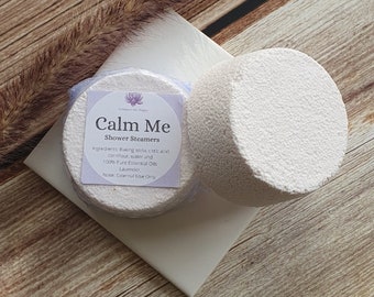 May include: Two white shower steamers with a textured surface, one round and one circular. The circular steamer has a label that reads "Calm Me Shower Steamers" with ingredients listed. The steamers rest on a white square and a wooden surface.