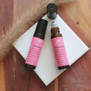 May include: Two essential oil roller bottles with pink labels that read "Passion". The bottles are brown with black caps. The labels list ingredients including lime and ylang-ylang essential oils. One cap is off, next to the bottles.