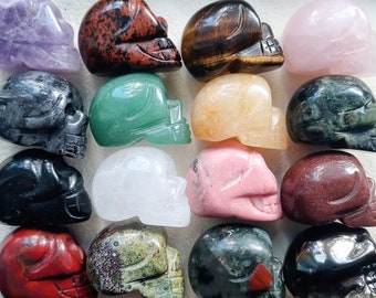 May include: A collection of carved gemstone skulls in various colors, including amethyst, red obsidian, tiger's eye, and rose quartz. The skulls are displayed in a grid pattern, showcasing their unique textures and hues. Each skull is approximately 1.5 inches in length.