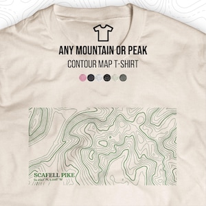 May include: A cream-coloured t-shirt displaying a green contour map of Scafell Pike, with coordinates. The text "ANY MOUNTAIN OR PEAK CONTOUR MAP T-SHIRT" is above the map. A small t-shirt icon is at the top.