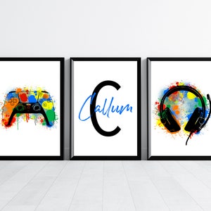 May include: Three framed prints with a black background and white border. The left print features a colourful paint splatter design with a black video game controller. The middle print features a large black letter C with the name Callum written in blue below. The right print features a colourful paint splatter design with a black headset.