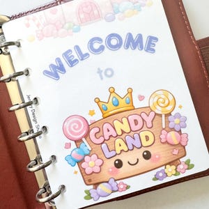 May include: A planner insert with a colorful "Welcome to Candy Land" design. The insert features a cartoon candy theme with lollipops, candies, and a smiling candy land sign. The insert is inside a brown leather planner.