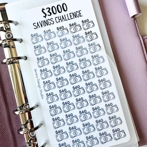 May include: A white plastic savings challenge tracker card with black text reading $3000 SAVINGS CHALLENGE and Jenavy Design Studio. The card features a grid of 50 small banknote icons, each labeled $60, inserted into a clear binder pocket.
