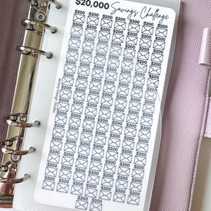 May include: A clear plastic savings challenge tracker with the text "$20,000 Savings Challenge" and envelope icons, each representing $200. The tracker is inside a binder. The text "Jenavy Design Studio" is also visible.