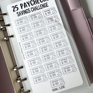 May include: A white savings challenge card with the text "25 PAYCHECKS SAVINGS CHALLENGE" and boxes to track savings. Each box is labeled "PAYCHECK $50" with a checkmark. The total savings amount is $1,250. The card is inside a clear plastic sleeve.
