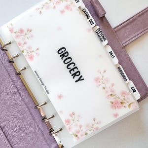 May include: A translucent plastic binder divider with a pink cherry blossom pattern and the text "GROCERY" printed in black. The divider features labeled tabs for "GROCERY," "EATING OUT," "PERSONAL," "BILLS," "HOUSEHOLD," and "CAR" inside a purple binder.