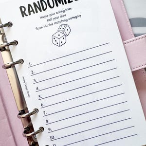 May include: A white planner insert with the word "RANDOMIZER" in bold black letters. Below the title are instructions and a dice graphic. The page has numbered lines for listing categories. The planner is in a pink binder.
