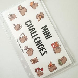 May include: Transparent plastic pouch containing a white insert with cartoon brown animals and food. The insert reads "Mini Challenges" in black text. The pouch has a zip closure and is designed for organisation.