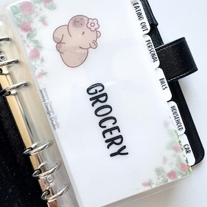 May include: A planner with a clear plastic cover and black binder. The "Grocery" divider features a cartoon hippo with a flower and floral accents. Additional dividers include "Eating Out," "Personal," "Bills," "Household," and "Car."