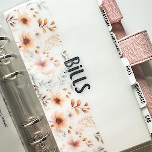 May include: A clear planner with floral design and the word "Bills" in black. The planner has tabs labeled "Debt", "Personal", "Emergency", "Bills", "Household", and "Car". The planner is held together with silver rings and has a pink cover.