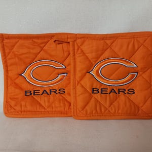 Pode incluir: Luvas de forno acolchoadas laranja com o logotipo dos Chicago Bears bordado em branco e azul marinho. A palavra "BEARS" é bordada abaixo do logotipo em cada luva. As luvas de forno têm uma argola para pendurar.