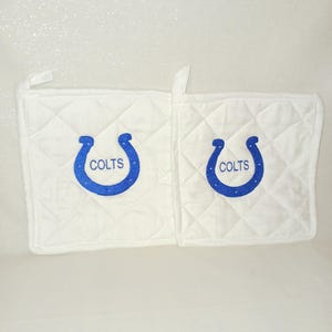 Puede incluir: Dos manoplas blancas acolchadas con un diseño de herradura azul y la palabra "COLTS". Cada manopla tiene un patrón de diamantes y un lazo para colgar.