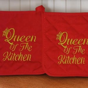 May include: Two square red quilted pot holders featuring gold embroidered text that reads Queen of the Kitchen with a small crown icon above the word Queen. Each pot holder has a fabric loop at the top corner for hanging purposes.