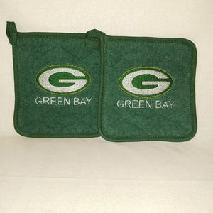 Op de afbeelding: Twee groene pannenlappen met de woorden "GREEN BAY" geborduurd onder een wit en groen "G" logo. De pannenlappen zijn vierkant met een ophanglus.