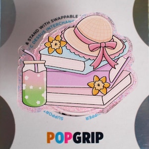 May include: A glittery acrylic phone grip topper featuring an illustration of two stacked books, a straw hat with a pink ribbon, two yellow daffodils, and a glass with a pink and green gradient drink with white flower patterns and a straw.