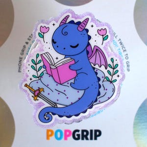 May include: A PopGrip featuring a blue cartoon dragon reading a pink book, with purple wings and horns. The design includes a sword, flowers, and the text "POPGRIP". The background is white with a glittery border.