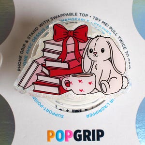May include: A PopGrip phone accessory featuring a stack of red books, a red bow, a white bunny, and a teacup with hearts. The accessory is white with glitter and has the words "PopGrip" in orange.