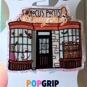 May include: A glittery phone grip featuring a Hocus Pocus Candy Shop illustration. The shop front has large windows displaying ghosts, broomsticks, and candy apples. The text Hocus Pocus Candy Shop is printed above the storefront. The base is a PopGrip.
