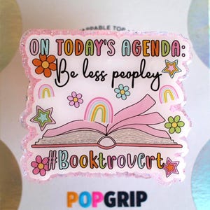May include: A PopGrip with a pink glitter border featuring the text "ON TODAY'S AGENDA: Be less peopley" in a whimsical font. The design includes an open book, rainbows, flowers, stars, and the hashtag #Booktrovert.