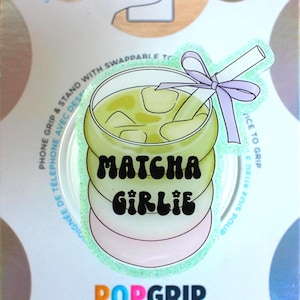 May include: A PopGrip phone grip featuring a matcha drink design with the words "Matcha Girlie". The grip has a light green drink with ice cubes, a straw, and a purple bow. The PopGrip is on a white background.