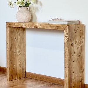 May include: A rustic wooden console table with a natural wood finish. The table has a rectangular top and two vertical supports. A vase with flowers and a book are on the table.