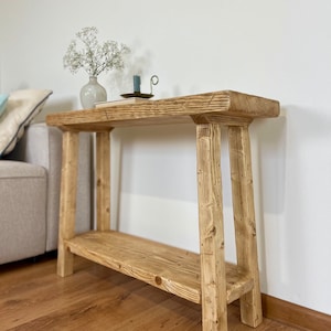 Könnte beinhalten: Ein rustikaler, zweistufiger Konsolentisch aus Holz mit natürlicher Oberfläche. Der Tisch hat ein unteres Regal und eine obere Ablage, die von stabilen Beinen getragen werden. Eine kleine Vase mit weißen Blumen und ein Kerzenhalter mit einer blauen Kerze stehen auf der oberen Ablage.