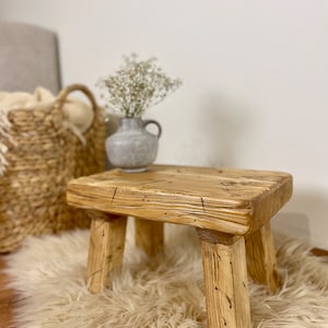 Könnte beinhalten: Ein rustikaler Holzhocker mit naturbelassenem Finish und vier Beinen. Der Hocker steht auf einem flauschigen beigefarbenen Teppich.