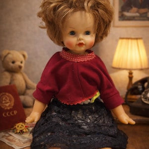 Peut inclure: Une poupée vintage avec un haut rouge et une jupe en dentelle noire. La poupée a une perruque blonde et est assise sur une surface en bois. Un ours en peluche et une image encadrée sont en arrière-plan.