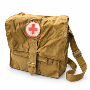 Op de afbeelding: Een beige schoudertas van canvas met een opvallende witte ronde patch met een rood kruis op de voorflap. De tas is voorzien van een enkele verticale riem met een metalen gespsluiting en een verstelbare schouderband met een metalen schuif.