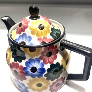 May include: A white ceramic teapot featuring a repeating pattern of stylized flowers in shades of yellow, blue, red, and dark green. The teapot includes a matching lid with a black knob, a black spout, and a black angular handle.