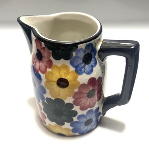 Può includere: Una lattiera in ceramica con base bianca decorata con un motivo ripetitivo di fiori multicolori nelle tonalità del blu, giallo, rosa e verde scuro. La lattiera presenta un manico blu scuro e un bordo blu scuro attorno al beccuccio.