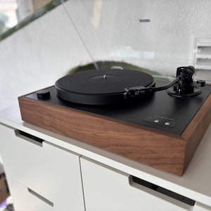 May include: A black turntable with a wooden base and a clear plastic cover. The turntable features a black platter, tonearm, and control knob. The wooden base has a rich, dark brown finish. The turntable is placed on a white surface.