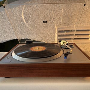 Op de afbeelding: Een Technics platenspeler met een donkere houten basis en een zilverkleurige metalen bovenplaat. Het apparaat is voorzien van een doorzichtige kunststof stofkap, een zwarte vinylplaat op het draaiplateau en een zilverkleurige toonarm.
