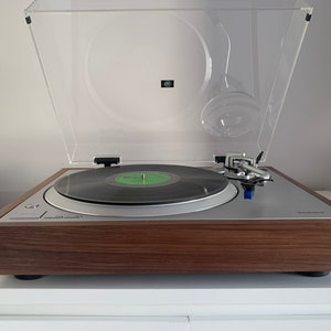 Op de afbeelding: Een zilveren draaitafel met een zwarte vinylplaat op de draaiplateau. De draaitafel heeft een transparante stofkap en een zilveren toonarm. De draaitafel staat op een houten basis.