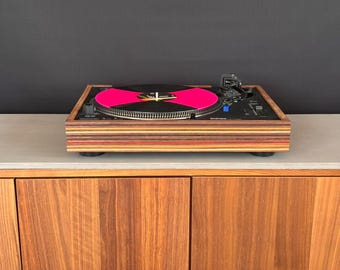 Puede incluir: Un tocadiscos con base negra y un marco de madera a rayas de varias capas. El plato sostiene un disco de vinilo negro con una etiqueta central de color rosa intenso. El dispositivo incluye un brazo fonocaptor negro, un control de tono y botones.