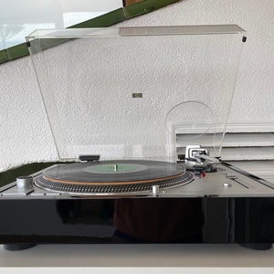 May include: A black turntable with a clear dust cover and a silver tone arm. The turntable is on a white surface.