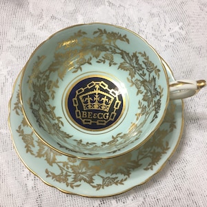 May include: A light blue teacup and saucer with a gold floral pattern and a blue and gold crest with the text "BE & CO" in the centre.