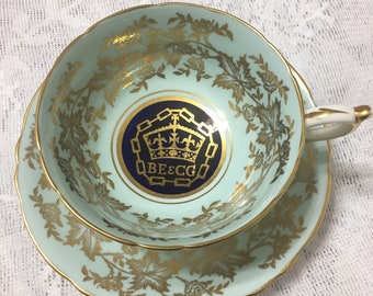 Könnte beinhalten: Eine hellblaue Teetasse und Untertasse mit einem goldenen Blumenmuster und einem blauen und goldenen Wappen mit dem Text "BE & CO" in der Mitte.