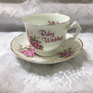 Può includere: Una tazza da tè e un piattino bianchi con un motivo di rose rosa. La tazza da tè ha un bordo dorato e le parole "Ruby Wedding" scritte in rosa sul lato.