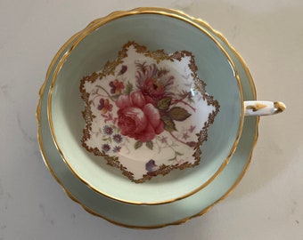 Peut inclure: Un service à thé vintage composé d'une tasse et d'une soucoupe. La tasse et la soucoupe sont vert pâle avec des bordures dorées. L'intérieur de la tasse présente un motif floral avec une grande rose rose et de petites fleurs violettes.