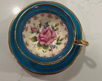 May include: A vintage teacup and saucer set with a floral design. The cup and saucer are both a vibrant turquoise blue with gold trim. The floral design features a pink rose with green leaves and blue flowers.