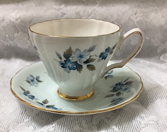 May include: A white teacup and saucer with a blue and green floral pattern. The cup and saucer have a gold trim.