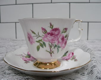 Puede incluir: Una taza de té y un platillo blancos con un patrón floral de rosas rosas. La taza tiene un borde dorado y una banda dorada alrededor de la base. El platillo tiene un borde festoneado.