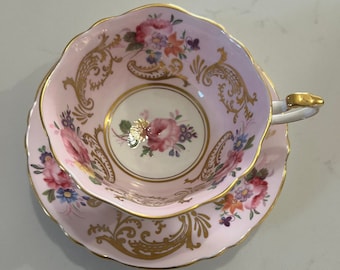 May include: A pink teacup and saucer set with a floral pattern and gold accents. The cup and saucer have a delicate, scalloped edge.